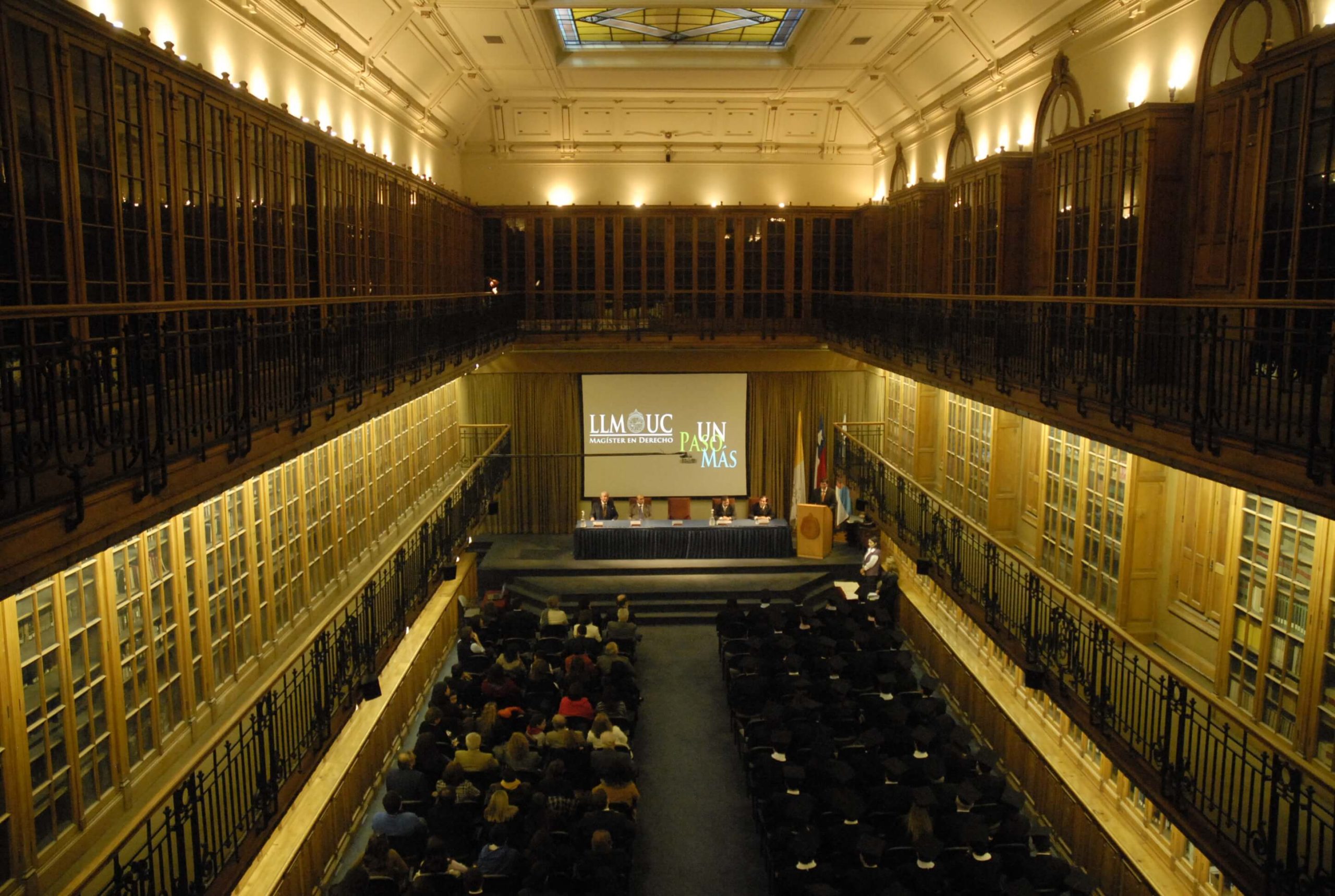 Gran salón de un edificio, con personas sentadas en sillas y un escenario